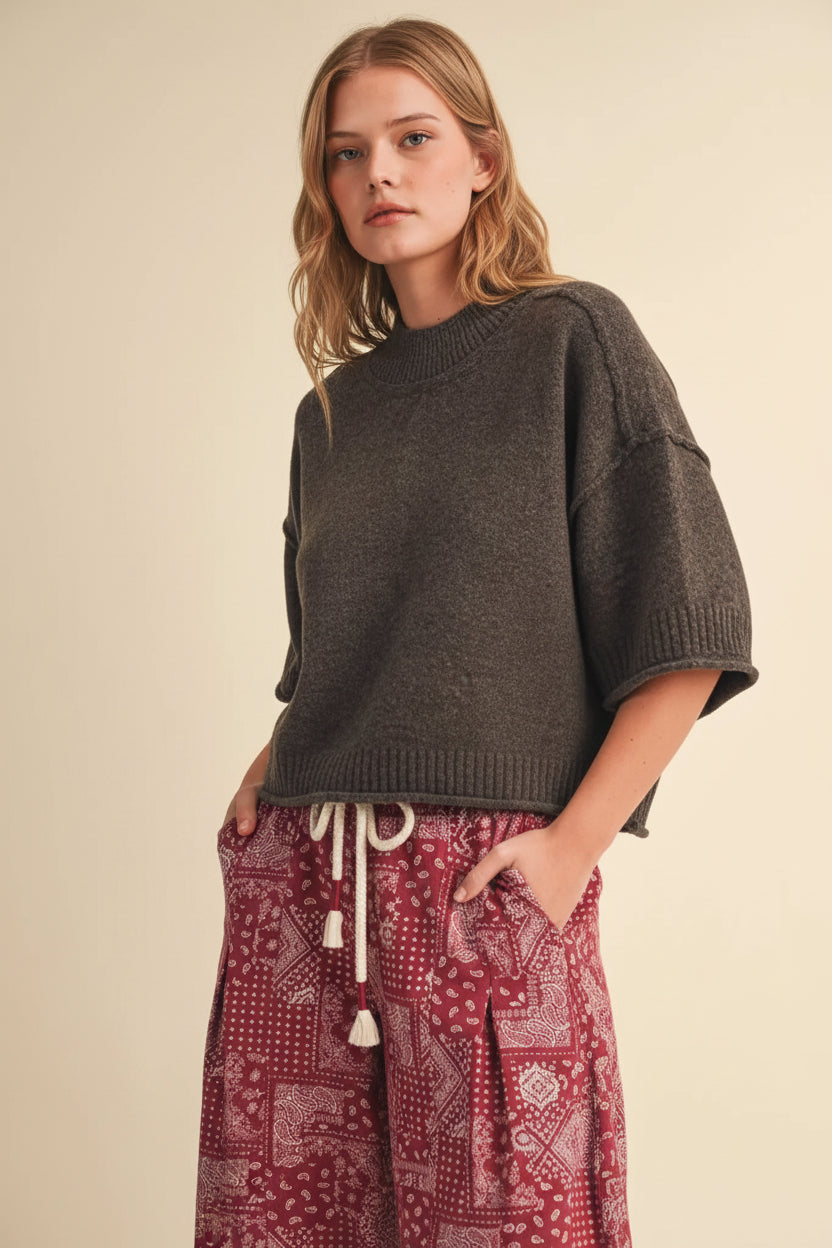 A woman modeling an oversized dark brown knit sweater with wide short sleeves and a cropped hem, paired with red patterned pants, against a plain beige studio background.